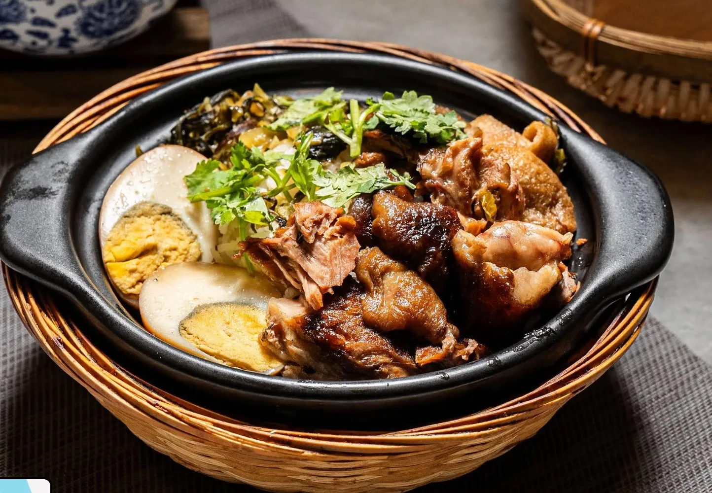 Braised meat with halved braised eggs, pickled greens and coriander in a black claypot, set in a woven bamboo basket