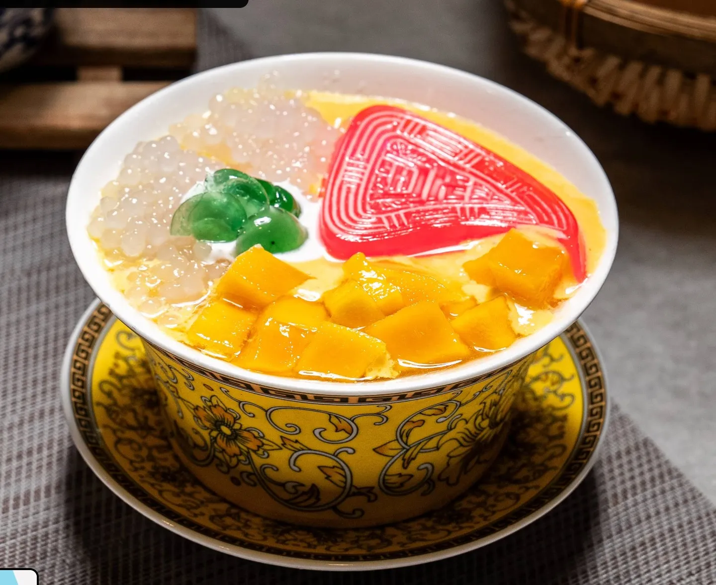 A yellow patterned bowl of golden sweet soup topped with sago pearls, diced mango and a red patterned kueh, on a matching plate